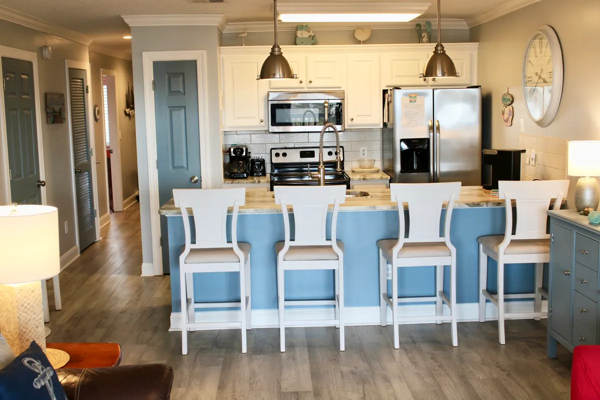 This image showcases a modern kitchen with a large island, sleek appliances, bar stools, and colorful decor, clearly designed for socializing and entertaining. The island invites guests to gather for drinks or casual bites, while the vibrant decor adds a sense of celebration and togetherness—matching the narrative of friends cooking and dining together.