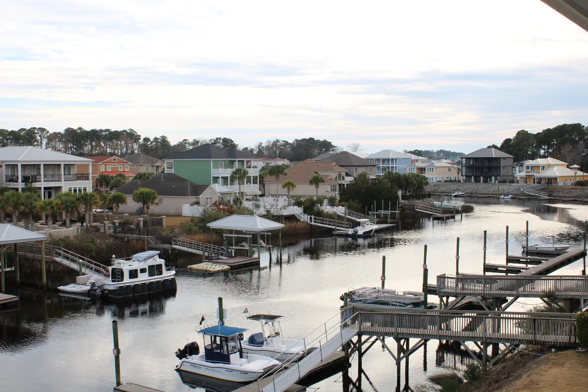 A wide panoramic view of the Intercoastal Waterway featuring sparkling blue water, boats drifting by, and lush palm trees lining the shore. Docks and waterfront homes complete the picturesque, resort-style scene.