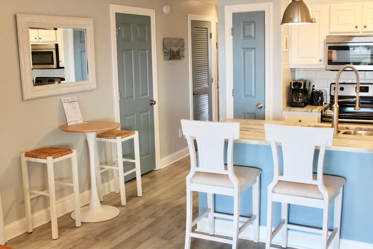 This image features a bright, welcoming kitchen area with bar stools around a kitchen island, kitchen appliances, and a framed mirror. Sunlight streams in, creating a connection between indoor comfort and the anticipation of heading outside for a day of exploration.