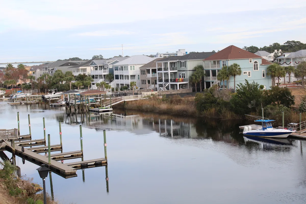 A scenic exterior shot featuring a tranquil waterway bordered by docks, boats, and lush vegetation. The image captures a peaceful setting ideal for waterfront relaxation, with houses visible along the shoreline and ample space for activities like boating or fishing.