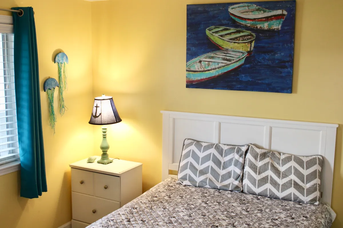 A cozy, cheerful sitting area accented by blue curtains, a colorful boat painting, nautical-themed lamp, and chevron-patterned pillows. The space is permeated with natural light, evoking a beachside ambiance and a sense of relaxation.