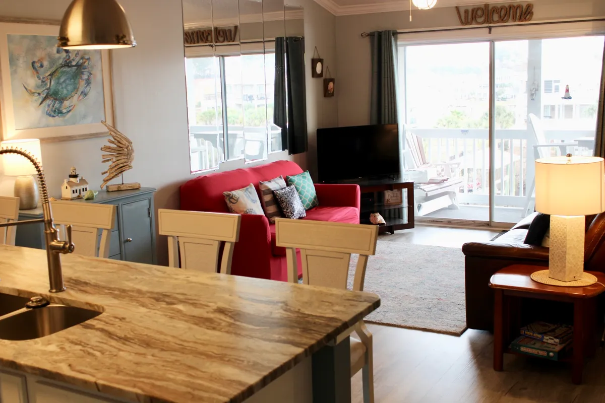 This image presents a wide view of the open-concept living area, with a striking red sofa, a kitchen island with bar stools, a welcoming sign, a television, and decorative art. The space feels cozy and social, with the kitchen and living space flowing together.