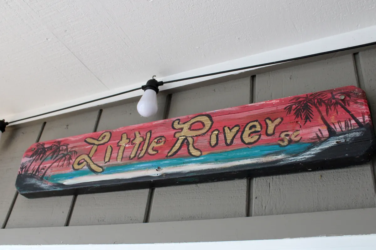 A vibrant, hand-painted wooden sign set against a colorful background, paired with a decorative light bulb that creates a welcoming atmosphere. The image evokes the laid-back, communal spirit of outdoor gatherings, perfect for family barbecues or evening get-togethers.