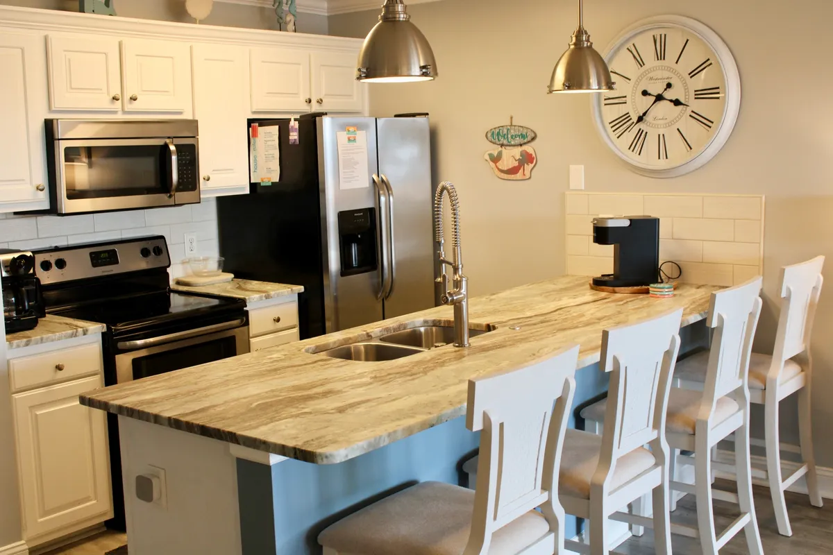 The image highlights a modern kitchen with granite countertops, stainless steel appliances, a large clock, and bar stools. Natural light streams in, and the space is arranged for both casual mornings and larger gatherings.