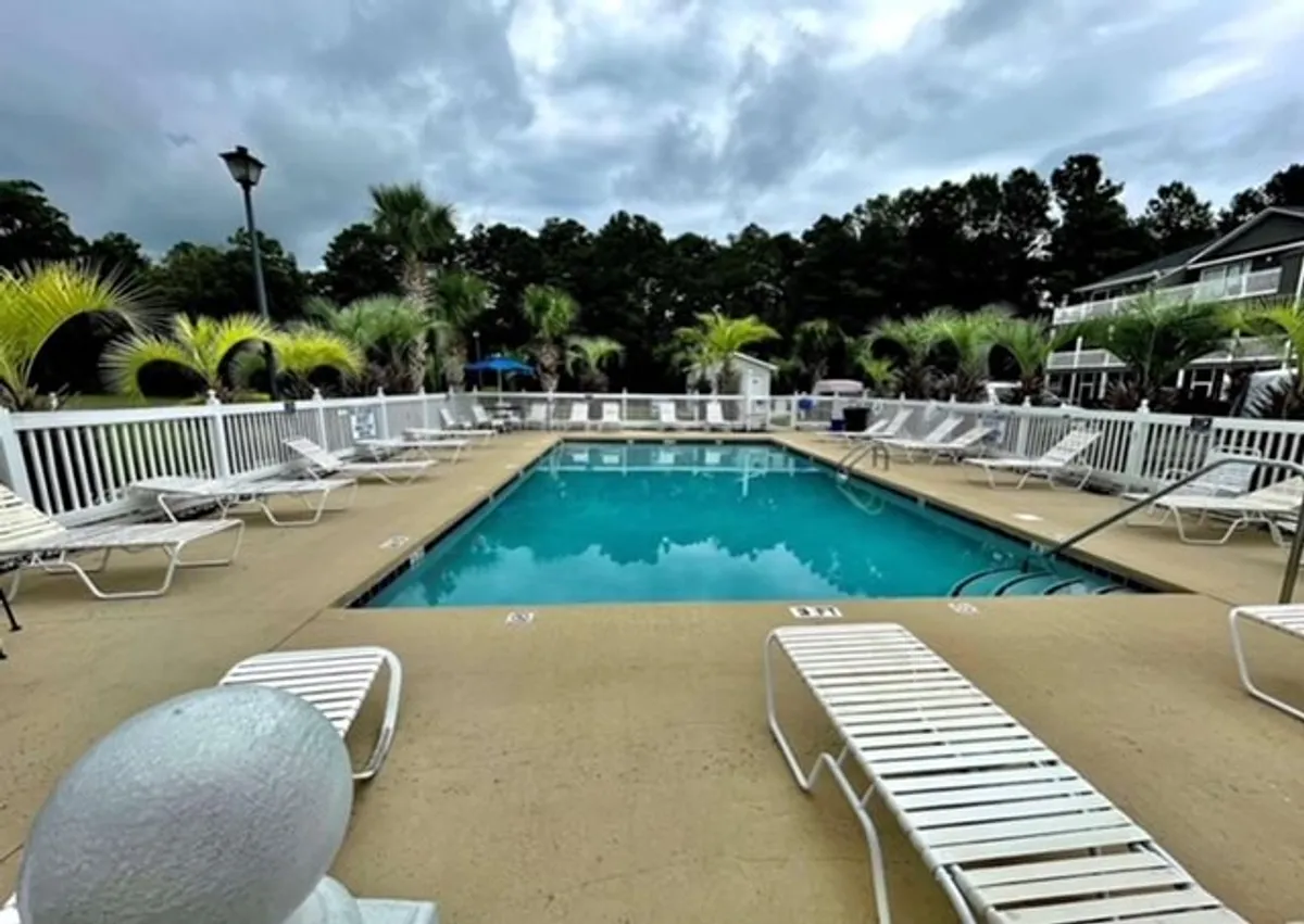 Another angle of a community swimming pool, featuring lounge chairs, palm trees, and inviting lighting fixtures. The setting is relaxed, with the pool flanked by greenery and a building in the background, suggesting an oasis-like retreat for both leisure and social gatherings.