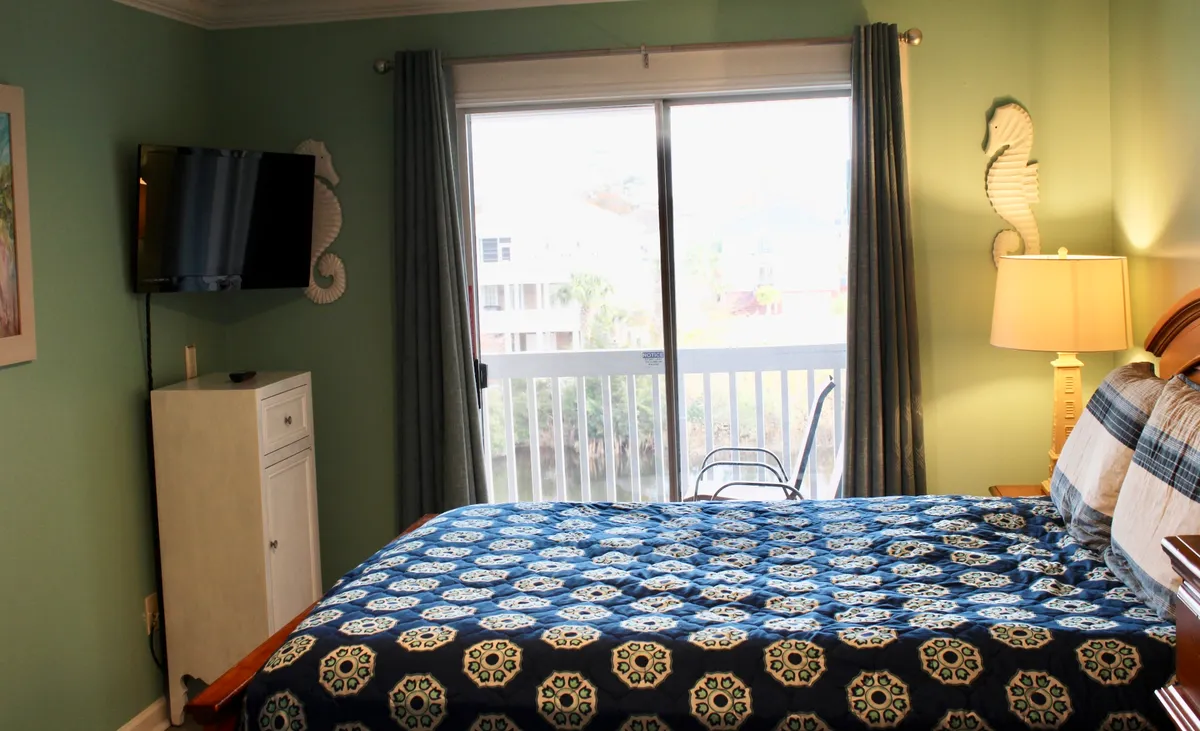 A bright bedroom featuring a bed with a blue patterned quilt, a large window offering scenic views, a bedside lamp, and a wall-mounted television. The space feels welcoming and designed for comfort, with natural light enhancing the room's appeal.