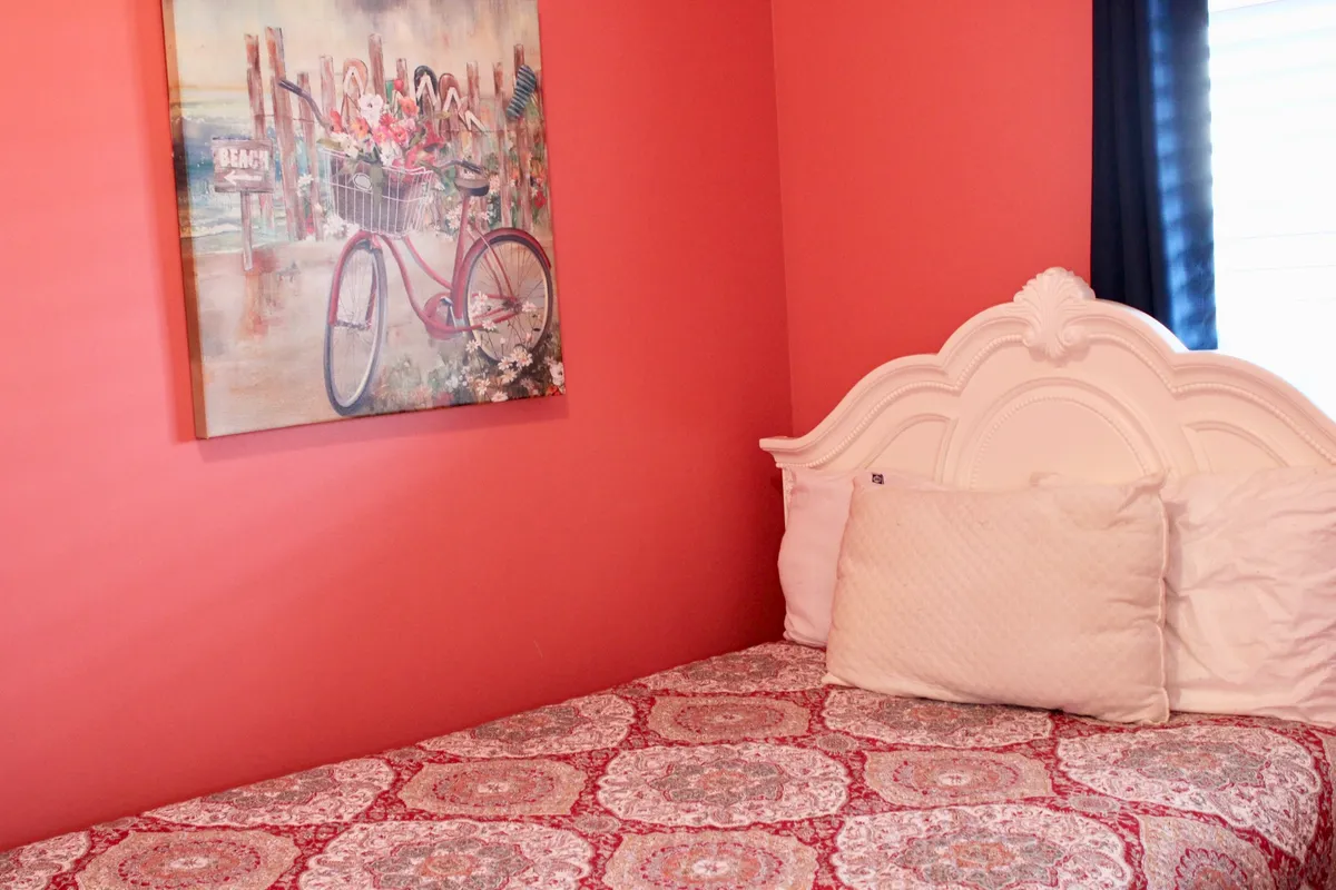 A serene bedroom scene showcasing a neatly made bed, pillow, and a painting above, bathed in soft light. The setting suggests quiet mornings, shared moments, and a tranquil atmosphere ideal for intimate escapes.